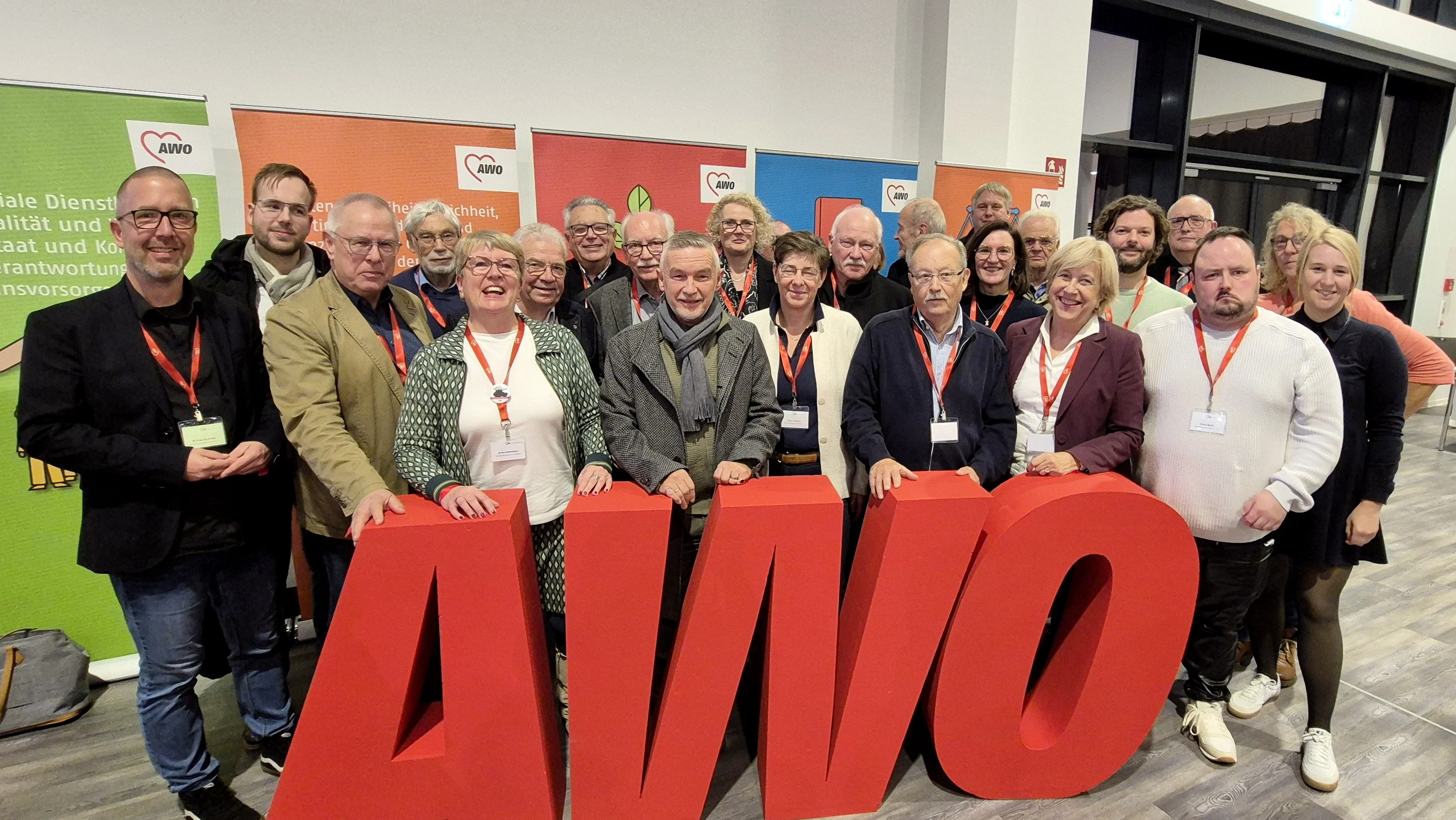 Auf dem Foto ist eine Gruppe von ungefähr zwanzig Erwachsenen zu sehen, die zusammensteht und in die Kamera blickt. Sie stehen hinter einem großen, roten 3D-Schriftzug, der die Buchstaben AWO (vermutlich für Arbeiterwohlfahrt) darstellt. Viele der Personen tragen Umhängebänder mit Namensschildern. Im Hintergrund sind Banner und Plakate zu erkennen, die ebenfalls das AWO-Logo (ein Herz) und Slogans zeigen. Auf dem Banner ganz links sind Teile eines Textes wie „soziale Dienstleistungen“ und „Qualität und Standards“ erkennbar. Der allgemeine Rahmen und die Kleidung der Personen (Mischung aus formell und leger) deuten auf eine Delegation oder Teilnehmergruppe bei einer Veranstaltung oder einem Kongress hin, wie es der Dateiname "Delegation Buko 2025" andeutet.