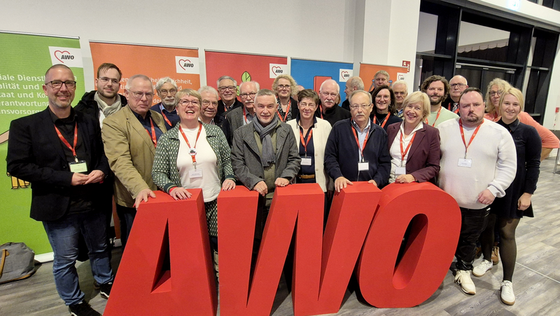  Auf dem Foto ist eine Gruppe von ungefähr zwanzig Erwachsenen zu sehen, die zusammensteht und in die Kamera blickt. Sie stehen hinter einem großen, roten 3D-Schriftzug, der die Buchstaben AWO (vermutlich für Arbeiterwohlfahrt) darstellt. Viele der Personen tragen Umhängebänder mit Namensschildern. Im Hintergrund sind Banner und Plakate zu erkennen, die ebenfalls das AWO-Logo (ein Herz) und Slogans zeigen. Auf dem Banner ganz links sind Teile eines Textes wie „soziale Dienstleistungen“ und „Qualität und Standards“ erkennbar. Der allgemeine Rahmen und die Kleidung der Personen (Mischung aus formell und leger) deuten auf eine Delegation oder Teilnehmergruppe bei einer Veranstaltung oder einem Kongress hin, wie es der Dateiname "Delegation Buko 2025" andeutet.