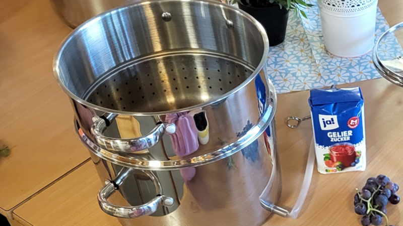 Auf dem Bild sind mehrere Kinder zu sehen, die gemeinsam dunkle Trauben vorbereiten. In der Mitte steht ein großer Edelstahl-Entsafter beziehungsweise Dampfentsafter; daneben liegen weitere Trauben und eine Packung Gelierzucker. 