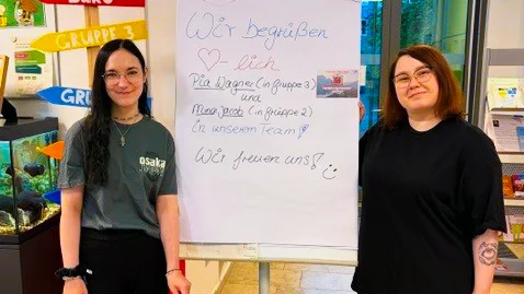 Zu sehen sind zwei junge Frauen in einem Eingangsbereich. Zwischen ihnen steht ein Flipchart mit einer handgeschriebenen Begrüßung für neue Auszubildende: „Wir begrüßen herzlich die Auszubildenden der Gruppe 3 und Mara … in unserem Team. Wir freuen uns!“ Im Vordergrund steht ein großes braunes Kiwi-Stofftier. Links weisen bunte Pfeilschilder unter anderem auf „Büro“ und „Gruppe 3“ hin; außerdem ist ein Aquarium zu sehen.