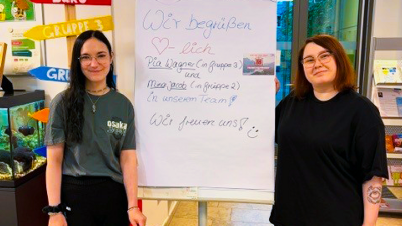 Zu sehen sind zwei junge Frauen in einem Eingangsbereich. Zwischen ihnen steht ein Flipchart mit einer handgeschriebenen Begrüßung für neue Auszubildende: „Wir begrüßen herzlich die Auszubildenden der Gruppe 3 und Mara … in unserem Team. Wir freuen uns!“ Im Vordergrund steht ein großes braunes Kiwi-Stofftier. Links weisen bunte Pfeilschilder unter anderem auf „Büro“ und „Gruppe 3“ hin; außerdem ist ein Aquarium zu sehen.