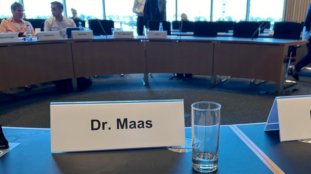 Das Foto zeigt einen Konferenzraum mit mehreren Personen, die an einem langen Tisch sitzen. Im Vordergrund befindet sich ein Namensschild mit der Aufschrift “Dr. Maas”, daneben steht ein leeres Glas. Im Hintergrund führen zwei Personen im Stehen ein Gespräch. Der Fokus des Bildes liegt offenbar auf dem Namensschild und einem Dokument davor.