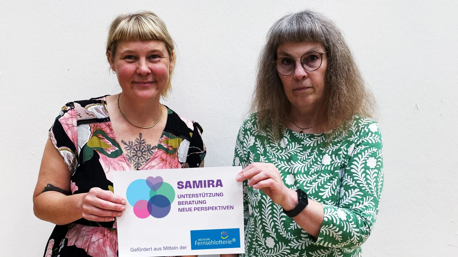 Zu sehen sind zwei Frauen vor einer weißen Wand. Sie halten gemeinsam ein Schild mit dem Logo und dem Namen „SAMIRA“ sowie dem Zusatz „Unterstützung · Beratung · Neue Perspektiven“ in die Kamera. Unten auf dem Schild steht, dass das Projekt aus Mitteln der Deutschen Fernsehlotterie gefördert wird.