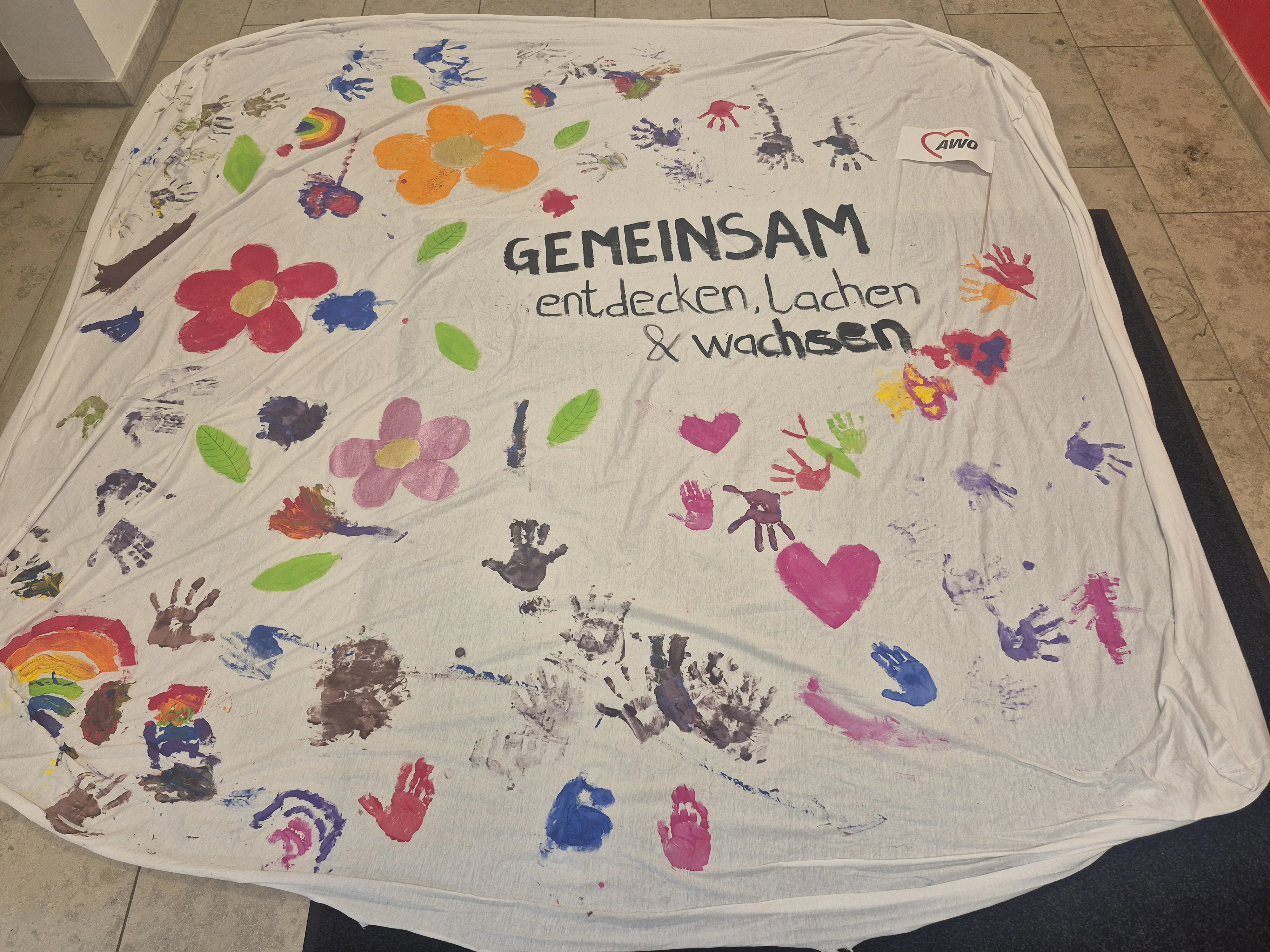 Auf dem Bild ist ein bemaltes, ausgebreitetes Tuch zu sehen, das offensichtlich in einer Aktion mit Kindern gestaltet wurde. In der Mitte steht der Schriftzug „GEMEINSAM entdecken, Lachen & wachsen“, daneben ist ein kleines Fähnchen mit dem AWO‑Herzlogo zu sehen. Rundherum sind bunte, kindlich gemalte Blumen, Blätter, Herzen, Regenbögen und viele farbige Handabdrücke in verschiedenen Größen verteilt. Das Tuch liegt auf einem Steinfußboden in einem Innenraum