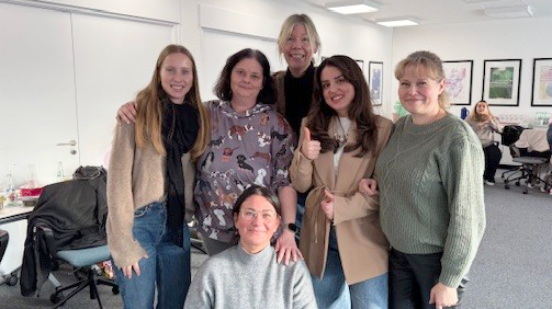 Auf dem Bild ist eine Gruppe von sechs Personen in einem Seminar‑ oder Büroraum zu sehen, fünf stehen nebeneinander, eine Person kniet vorne in der Mitte.