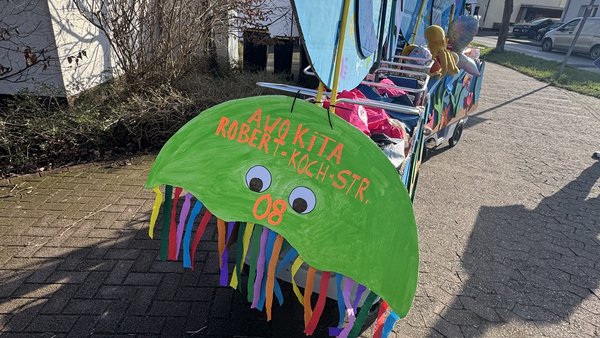 Auf dem Bild sieht man einen bunt dekorierten Bollerwagen, der wie ein Meerestierschiff gestaltet ist, mit großen, bemalten Fischen aus Pappe oder Holz an den Seiten. Vorne ist ein grünes „Quallen‑Schild“ mit Wackelaugen und bunten Papierstreifen sowie der Aufschrift „AWO KITA ROBERT‑KOCH‑STR. 08“.