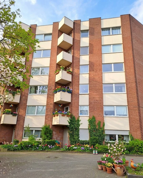 Auf dem Foto ist ein mehrstöckiges Wohnhaus zu sehen, mit mehreren Balkonen zur Vorderseite. Davor liegt eine gepflegte Rasenfläche mit Büschen und einigen großen Bäumen, am Rand verläuft ein niedriger Holzzaun entlang des Gehwegs.