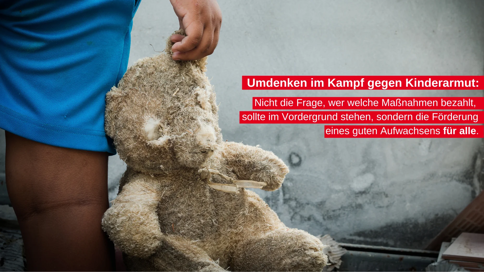 Das Foto zeigt eine Person in blauen Shorts, die neben einer Wand steht. Vor der Wand befindet sich ein großer Teddybär, dem die Gesichtszüge fehlen. An der Wand steht ein Text auf Deutsch: “Umdenken im Kampf gegen Kinderarmut: Nicht die Frage, wer welche Maßnahmen bezahlt, sollte im Vordergrund stehen, sondern die Förderung eines guten Aufwachsens für alle.”