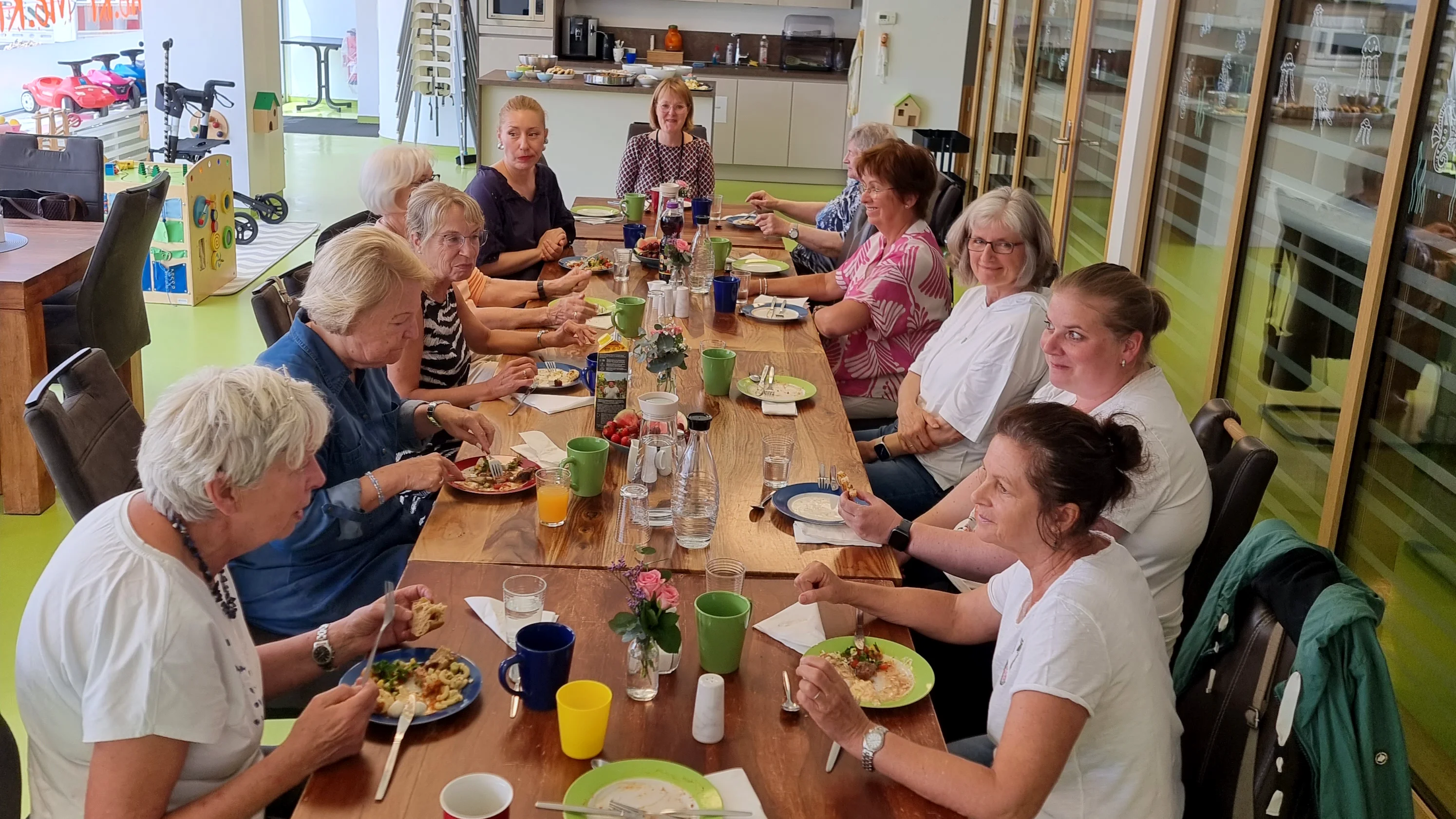 Auf dem Foto ist eine Gruppe von etwa 14 Frauen zu sehen, die gemeinsam an einem langen Holztisch in einem freundlich gestalteten Gemeinschaftsraum essen. Der Raum wirkt modern und einladend, mit großen Fenstern auf der rechten Seite und einer Küchenzeile im Hintergrund. Auf dem Tisch stehen bunte Tassen, Teller mit Essen, Gläser und Karaffen mit Wasser. Die Atmosphäre wirkt freundlich und gesellig. Im Hintergrund sind Spielgeräte und Hinweise auf eine AWO-Einrichtung sichtbar (z. B. das AWO-Logo auf einem Plakat an der Tür), was auf ein generationsübergreifendes oder soziales Angebot schließen lässt – möglicherweise ein gemeinsames Essen oder ein Treffpunkt für Ehrenamtliche, Seniorinnen oder Teilnehmerinnen eines Projekts.