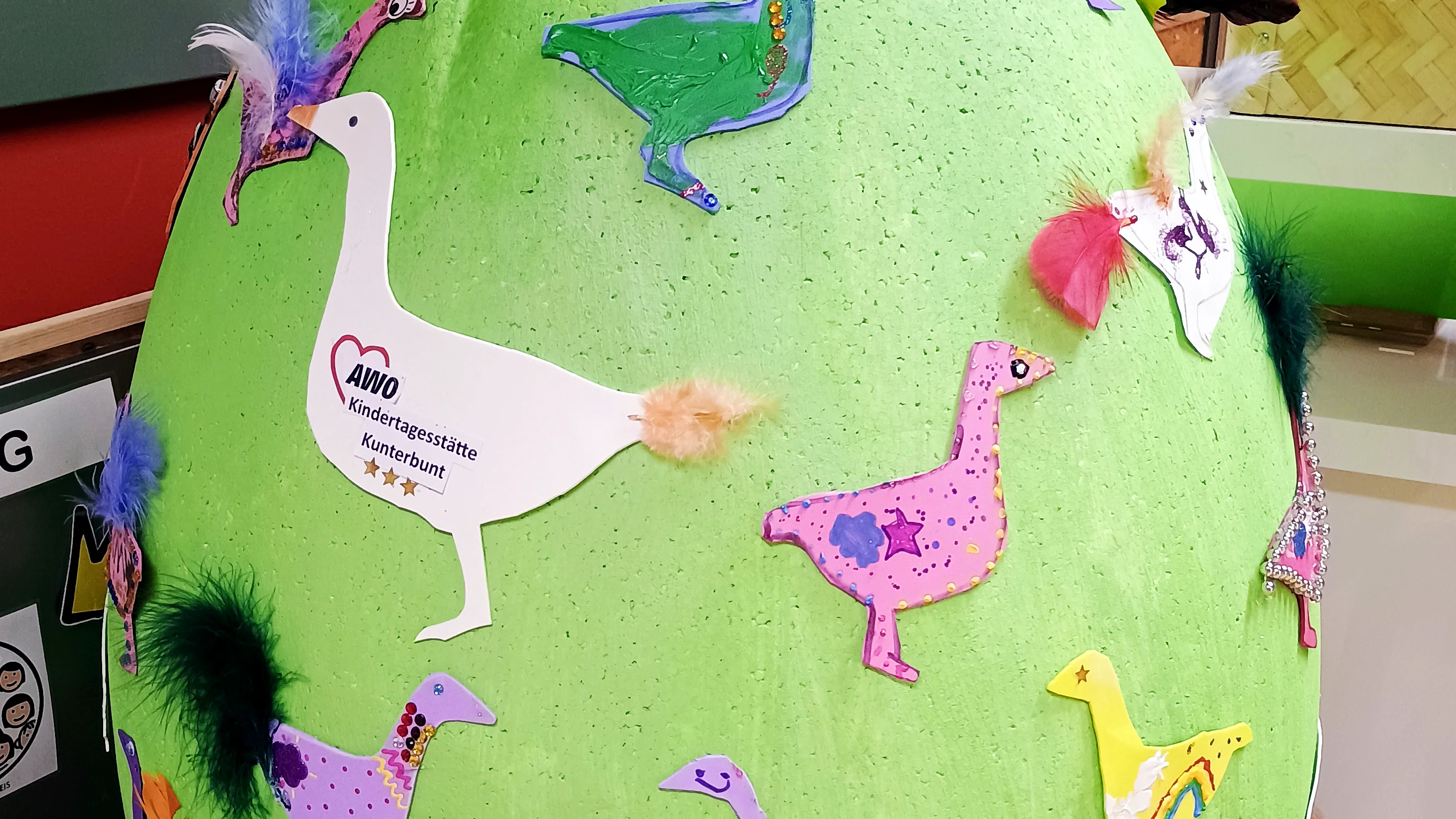 Das Foto zeigt ein großes, grün bemaltes Ei, das auf zwei kleinen Holzstühlen ruht. Das Ei ist mit zahlreichen bunten, ausgeschnittenen Gänsefiguren beklebt, die kreativ mit Farben, Federn und anderen Bastelmaterialien gestaltet wurden. Auf einer der weißen Gänse befindet sich ein Logo mit der Aufschrift „AWO Kindertagesstätte kunterbunt“, was darauf hindeutet, dass es sich um ein kreatives Bastelprojekt einer Kindertagesstätte handelt. Die Szene spielt sich vermutlich in einem Kindergarten oder einer ähnlichen Einrichtung ab.
