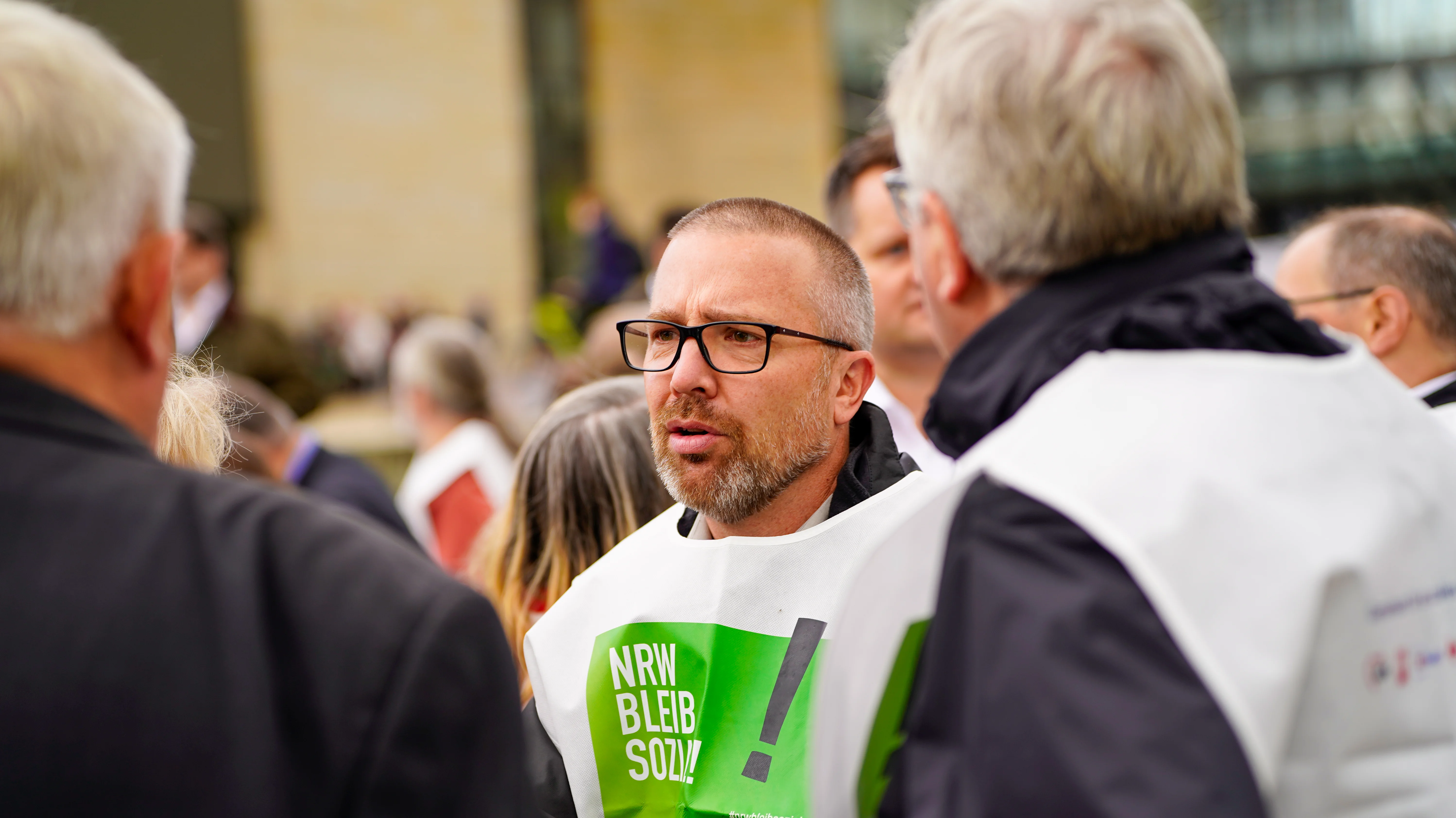 Das Foto zeigt eine Gruppe von Menschen von hinten. Im Vordergrund steht Michael Rosellen, der eine weiße Weste mit grünem Text trägt. Auf der Weste steht “NRW BLEIB SOZIAL" 