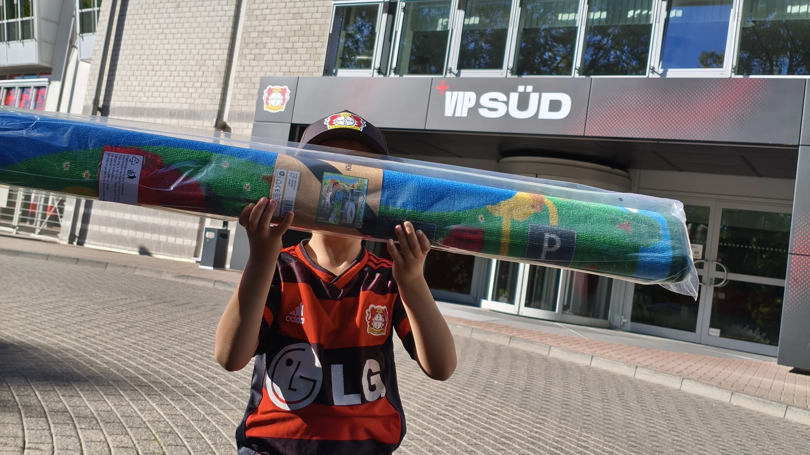 Auf dem Bild ist ein Kind vor einem Gebäude von Bayer 04 Leverkusen zu sehen. Das Kind trägt ein Leverkusen-Trikot und hält einen langen, verpackten Fan-Schal oder Teppich in den Händen. Im Hintergrund sind unter anderem die Beschriftungen „Bayer 04 Leverkusen“, „VIP Süd“ und ein Hinweis auf „Rückhalt“ zu erkennen.