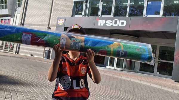 Auf dem Bild ist ein Kind vor einem Gebäude von Bayer 04 Leverkusen zu sehen. Das Kind trägt ein Leverkusen-Trikot und hält einen langen, verpackten Fan-Schal oder Teppich in den Händen. Im Hintergrund sind unter anderem die Beschriftungen „Bayer 04 Leverkusen“, „VIP Süd“ und ein Hinweis auf „Rückhalt“ zu erkennen.