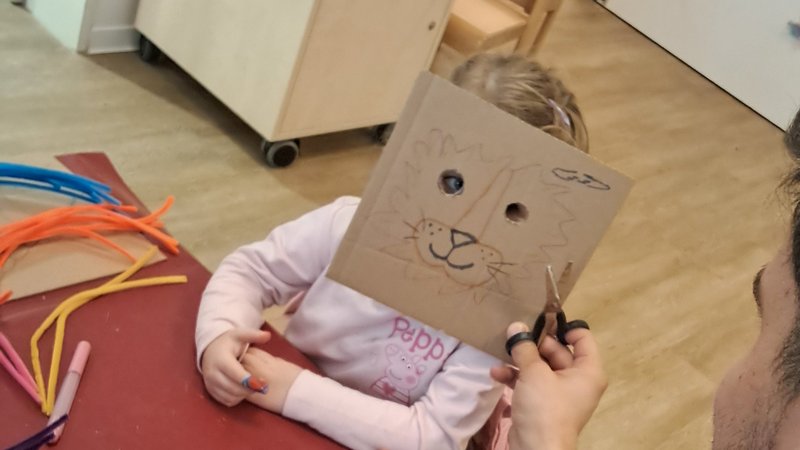 Das Bild zeigt eine Bastelszene in einem Kindergarten (erkennbar an dem Schild „Gruppe 2" mit bunten Vogelzeichnungen im Hintergrund). Ein Mann sitzt an einem rot abgedeckten Tisch und hält eine Schere sowie eine selbstgebastelte Pappmaske (Tier-/Hasengesicht) in den Händen. Gegenüber sitzt ein kleines Mädchen im rosa Pullover mit „Peppa"-Aufdruck. Auf dem Tisch liegen Stifte und bunte Pfeifenreiniger als Bastelmaterial. Es wirkt wie ein gemeinsamer Kreativ-/Bastelnachmittag in einer Kita-Einrichtung.