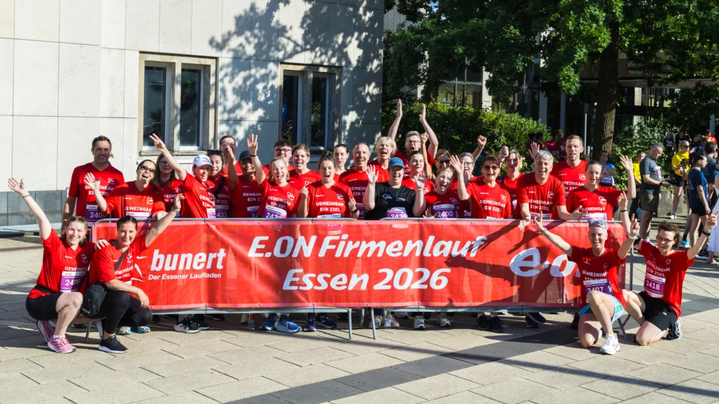 Auf dem Bild ist eine große Gruppe von Läuferinnen und Läufern in roten Shirts zu sehen, die fröhlich in die Kamera schauen und die Arme hochheben. Vor ihnen hängt ein rotes Banner mit der Aufschrift „E.ON Firmenlauf Essen 2026“ sowie weiteren Logos und Texten, das auf einen Firmenlauf in Essen hinweist.