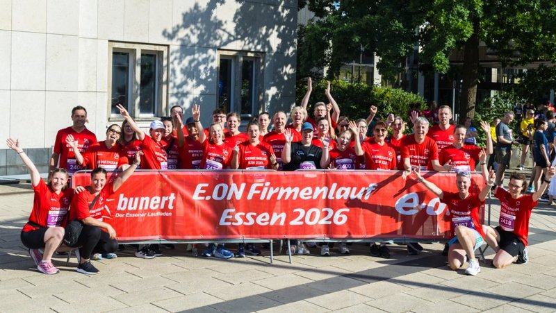 Auf dem Bild ist eine große Gruppe von Läuferinnen und Läufern in roten Shirts zu sehen, die fröhlich in die Kamera schauen und die Arme hochheben. Vor ihnen hängt ein rotes Banner mit der Aufschrift „E.ON Firmenlauf Essen 2026“ sowie weiteren Logos und Texten, das auf einen Firmenlauf in Essen hinweist.