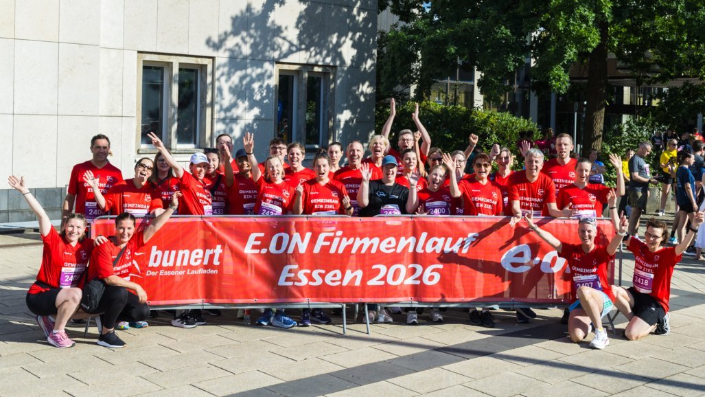 Auf dem Bild ist eine große Gruppe von Läuferinnen und Läufern in roten Shirts zu sehen, die fröhlich in die Kamera schauen und die Arme hochheben. Vor ihnen hängt ein rotes Banner mit der Aufschrift „E.ON Firmenlauf Essen 2026“ sowie weiteren Logos und Texten, das auf einen Firmenlauf in Essen hinweist.