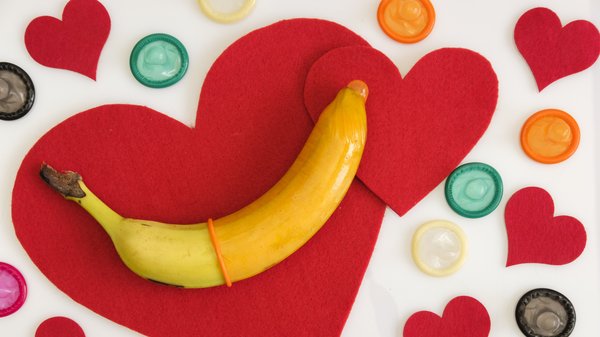 Auf dem Bild liegt eine Banane auf großen roten Filz-Herzformen, umgeben von mehreren bunten Kondomen, die kreisförmig um das Motiv verteilt sind. Die Szene symbolisiert offensichtlich das Thema Sexualität, Verhütung und „Safer Sex“ in einem verspielten, leicht humorvollen Stil.
