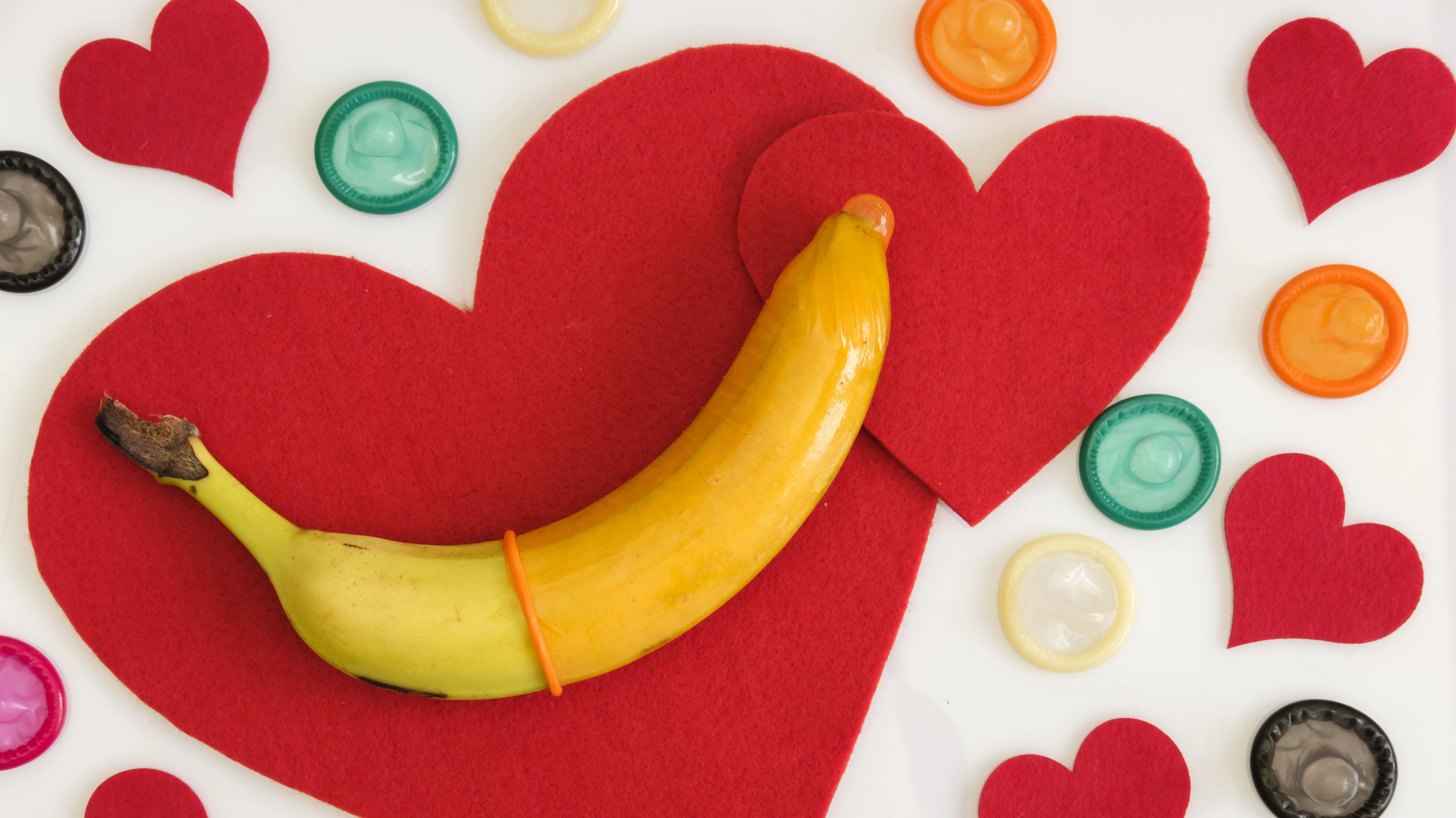 Auf dem Bild liegt eine Banane auf großen roten Filz-Herzformen, umgeben von mehreren bunten Kondomen, die kreisförmig um das Motiv verteilt sind. Die Szene symbolisiert offensichtlich das Thema Sexualität, Verhütung und „Safer Sex“ in einem verspielten, leicht humorvollen Stil.