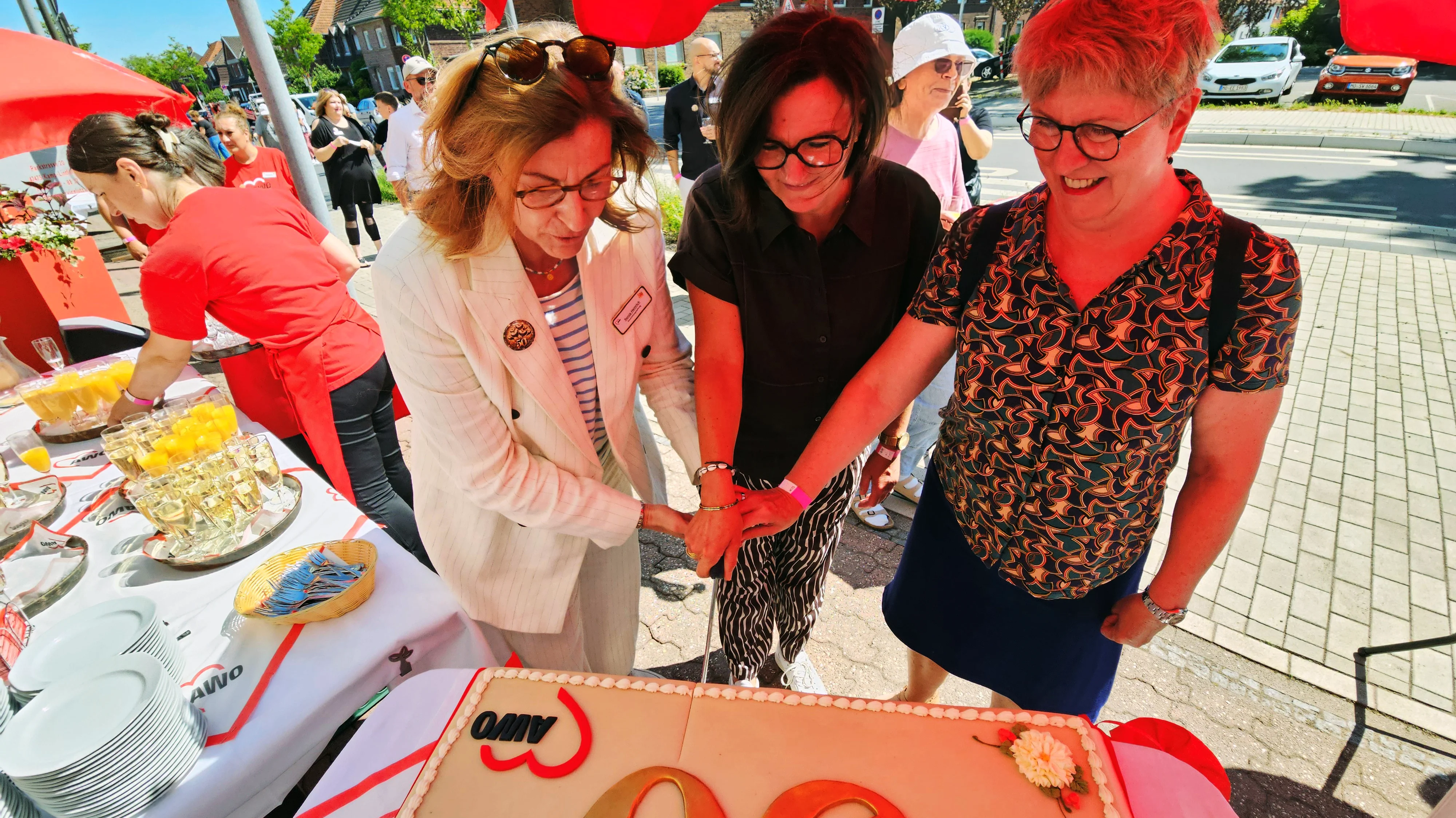 Auf dem Bild ist eine Feier im Freien zu sehen, bei der drei Frauen gemeinsam eine große Jubiläumstorte anschneiden. Unter einem roten Sonnenschirm stehen die drei im Vordergrund an einem festlich gedeckten Tisch mit einer rechteckigen Torte, auf der „50“ und das AWO‑Herzlogo zu erkennen sind. Im Hintergrund sind weitere Gäste, eine Frau im roten AWO‑Shirt an einem Getränketisch sowie eine Straße mit parkenden Autos und Häusern zu sehen.