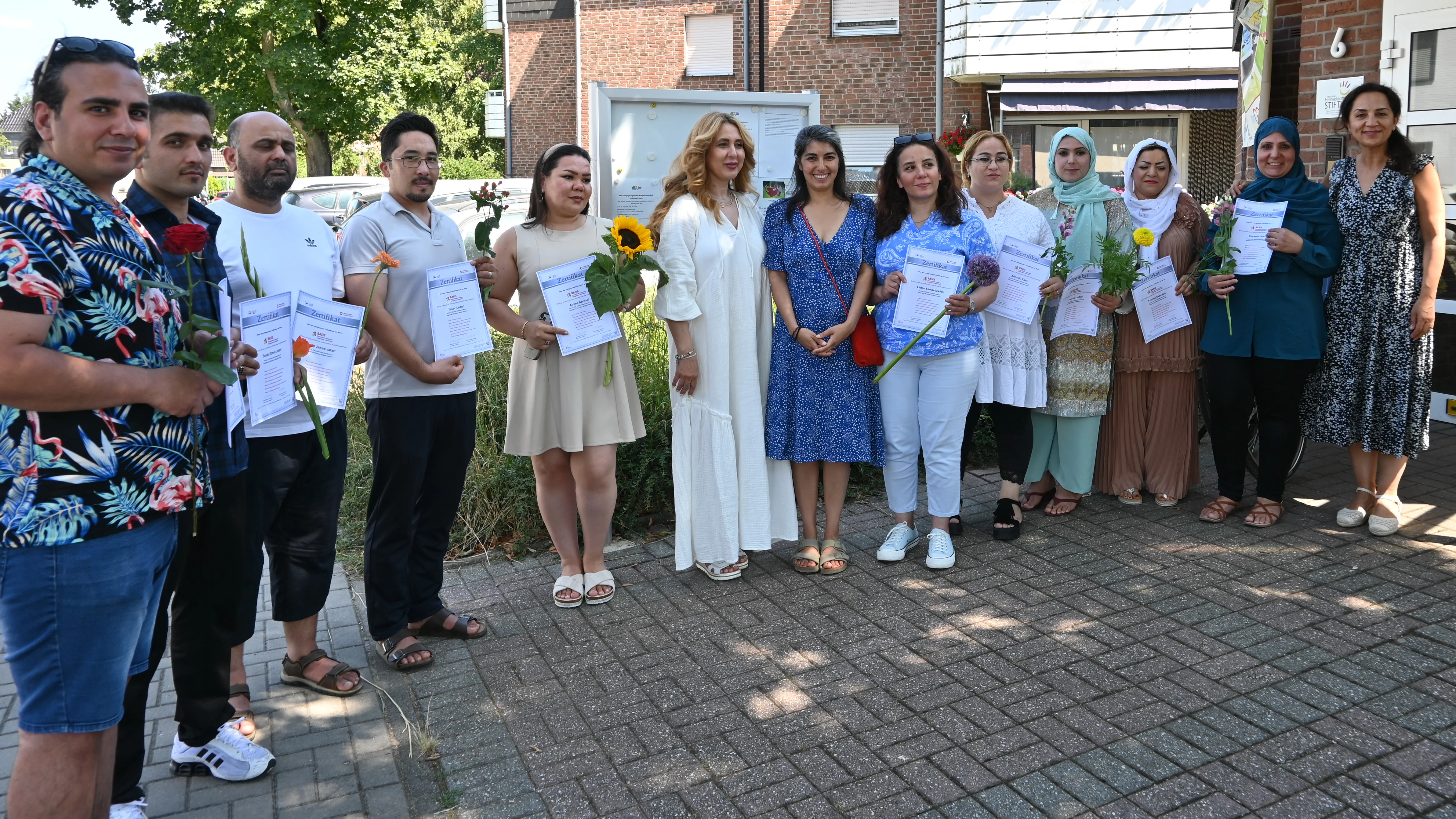 Teilnehmende eines Bildungs- und Qualifizierungsprogramms präsentieren ihre Zertifikate und Blumen vor einem Wohn- und Gemeinschaftsgebäude. Das Gruppenfoto wurde im Freien bei sonnigem Wetter aufgenommen und dokumentiert den erfolgreichen Abschluss der Maßnahme.