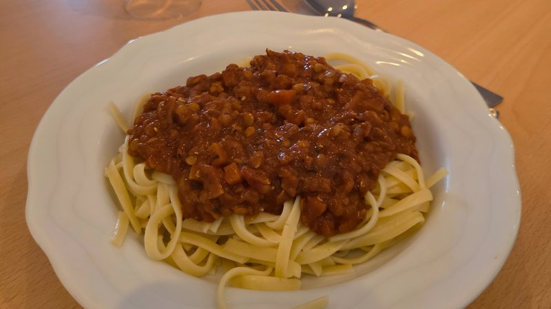Auf dem Bild steht ein Teller mit Pasta und Linsenbolognese im Vordergrund auf einem gedeckten Tisch, im Hintergrund sitzen mehrere Personen an einer langen Tafel mit Wasserflaschen und Deko.