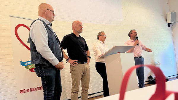 Auf dem Foto ist eine Veranstaltung der AWO (Arbeiterwohlfahrt) im Rhein-Kreis Neuss zu sehen. Vier Männer stehen vorne im Raum, einer von ihnen spricht gerade mit einem Mikrofon. Es scheint sich um eine offizielle Rede oder Ehrung zu handeln – möglicherweise im Rahmen einer Preisverleihung oder Auszeichnung. Im Vordergrund ist unscharf ein kleines Objekt auf einem Tisch zu erkennen – vermutlich eine dekorative Auszeichnung oder Verdienstmedaille mit rotem Band in Herzform. Im Hintergrund steht ein Roll-Up mit dem AWO-Logo und dem Slogan „ECHT ENGAGIERT“, daneben der Schriftzug „AWO im Rhein-Kreis Neuss“. Die Atmosphäre wirkt feierlich und offiziell.