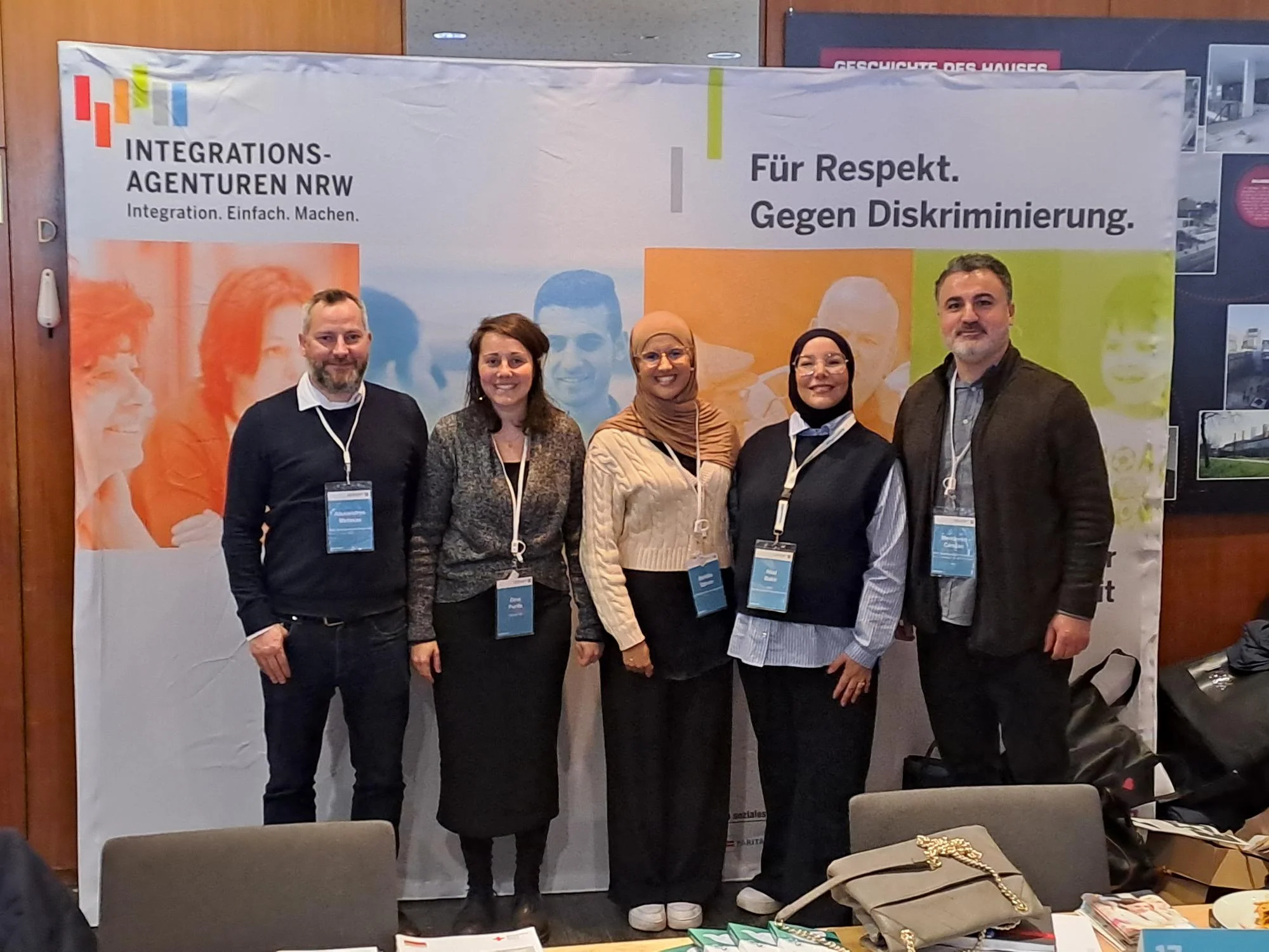 Das Foto zeigt eine Gruppe von fünf Personen, die vor einem Banner der Integrationsagenturen NRW stehen. Das Banner enthält die Slogans "Integration. Einfach. Machen." und "Für Respekt. Gegen Diskriminierung.". Die Personen tragen Namensschilder und sind Teilnehmende des 10. Integrationskongresses des Landes NRW. Im Vordergrund sind ein Tisch mit Broschüren, Unterlagen und Taschen zu sehen, was auf einen Informationsstand oder eine Ausstellung im Rahmen des Kongresses hindeutet. Die Atmosphäre wirkt professionell und engagiert, passend zum Thema Integration und Chancengleichheit.