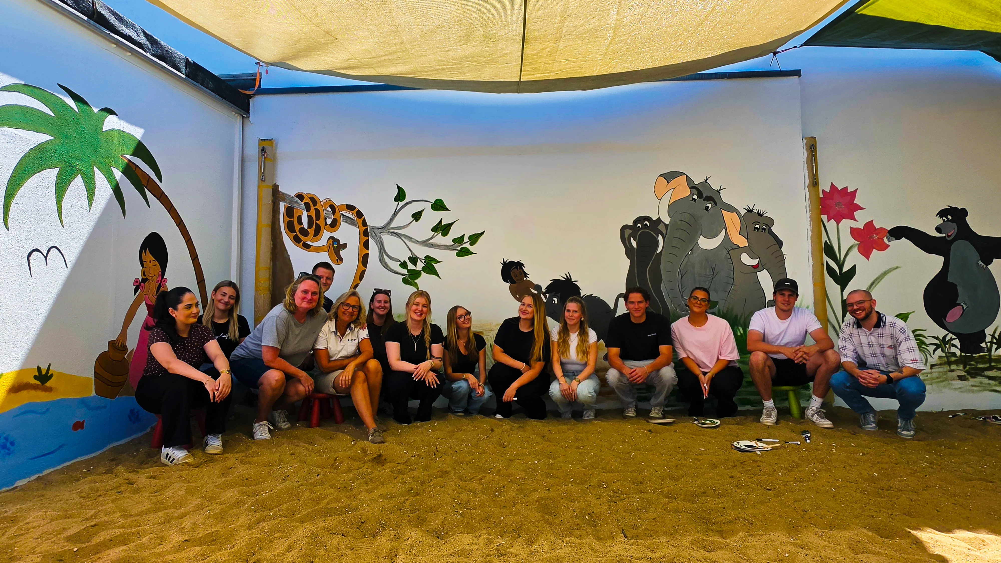 Auf dem Foto sieht man eine Gruppe von Menschen, die im Halbkreis auf kleinen Hockern oder direkt im Sand sitzen und in die Kamera lächeln. Im Hintergrund ist eine bunt bemalte Wand zu sehen, die offenbar eine Dschungel‑/„Dschungelbuch“-Szenerie zeigt: links ein Palmenstrand mit Wasser, daneben eine gezeichnete Figur und eine Schlange, in der Mitte mehrere gezeichnete Kinder oder Tiere, rechts eine Gruppe grau gemalter Elefanten sowie eine schwarze Bärenfigur mit einer großen roten Blume. Der Boden besteht aus Sand, über der Gruppe hängt ein Sonnensegel.