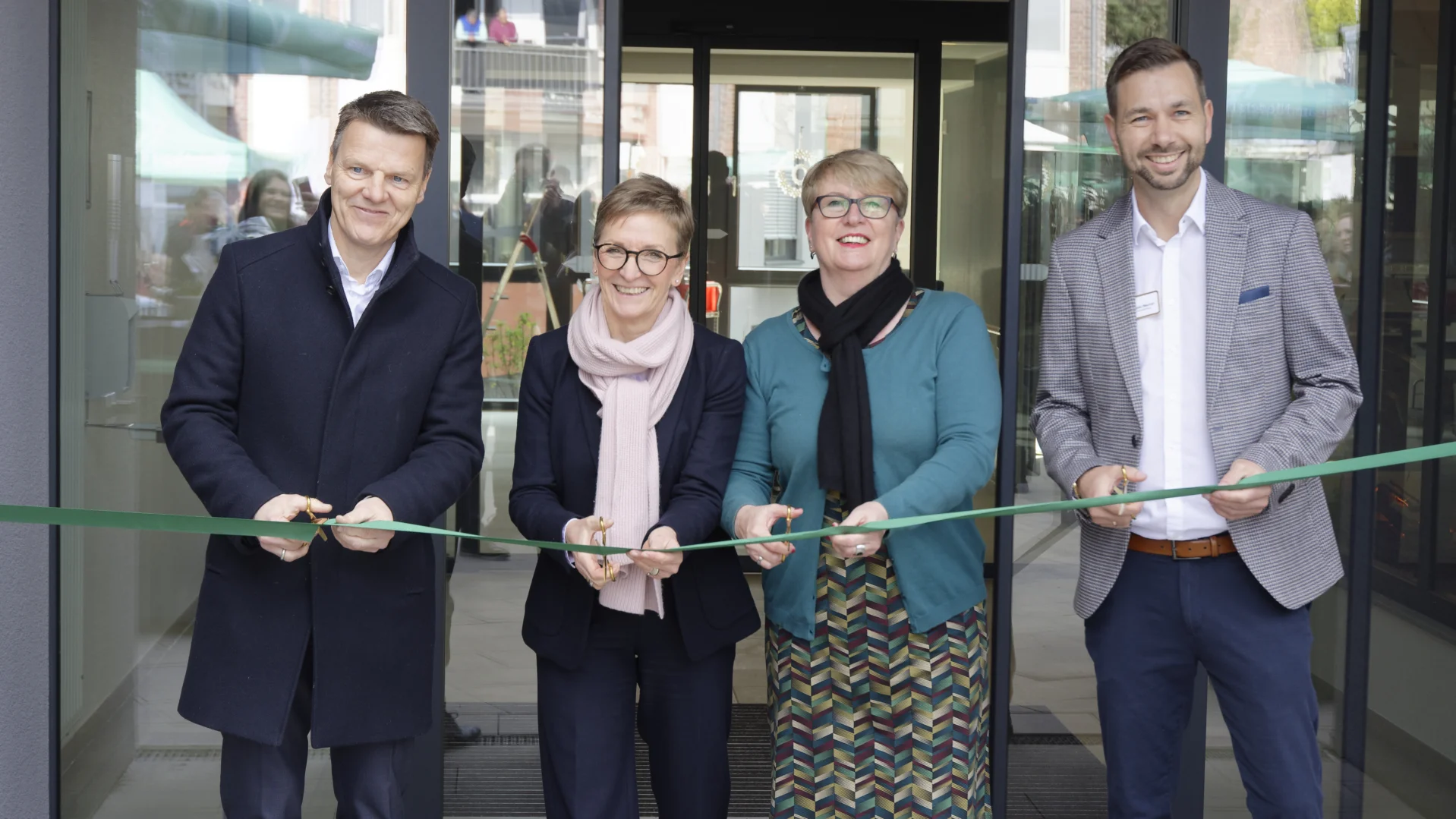 Auf dem Foto sieht es so aus, als ob eine feierliche Eröffnung stattfindet. Vier Personen stehen vor einem Gebäude und schneiden gemeinsam ein grünes Band durch – eine klassische Szene für eine Einweihungszeremonie. Alle scheinen gut gelaunt zu sein und lächeln in die Kamera.