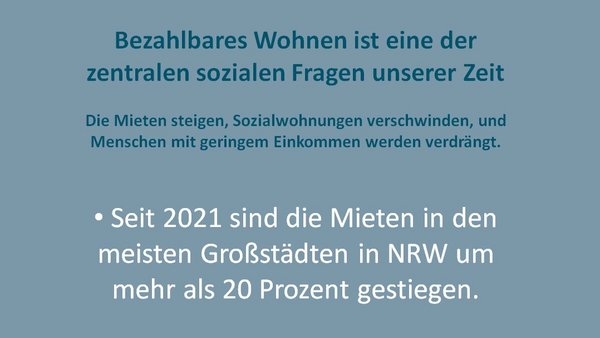 Das Foto zeigt ein Plakat mit dem Titel "Bezahlbares Wohnen ist eine der zentralen sozialen Fragen unserer Zeit". Es werden drei Hauptaussagen in Form von Bullet Points präsentiert: Seit 2021 sind die Mieten in den meisten Großstädten in Nordrhein-Westfalen (NRW) um mehr als 20 Prozent gestiegen. Der Bestand an Sozialwohnungen in NRW ist von 1,3 Millionen (1990) auf aktuell 422.000 gesunken. 2024 wurden in NRW 122.170 wohnungslose Menschen offiziell erfasst. Unterhalb des Textes befindet sich das Logo der Organisation "Wir wollen wohnen" und die zugehörige Webseite "www.wir-wollen-wohnen-nrw.de".