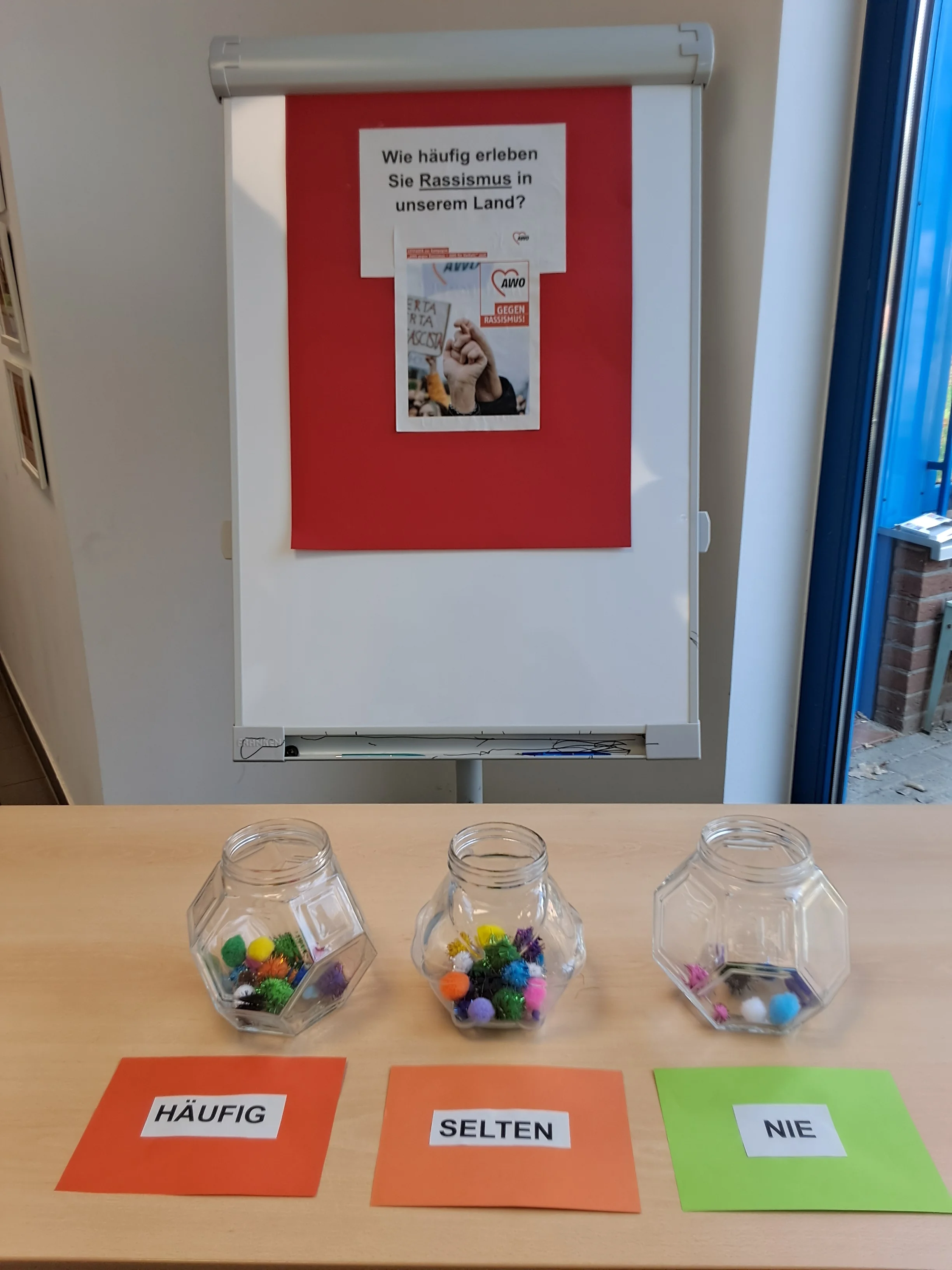 Auf dem Foto steht ein aufgestelltes Flipchart mit einem roten Plakat und der Frage: „Wie häufig erleben Sie Rassismus in unserem Land?“. Davor steht ein Tisch mit drei durchsichtigen Gläsern, in die bunte Pompons gelegt wurden. Vor jedem Glas liegt ein farbiges Blatt mit den Antwortmöglichkeiten „HÄUFIG“, „SELTEN“ und „NIE“. In den Gläsern bei „HÄUFIG“ und „SELTEN“ liegen deutlich mehr Pompons als im Glas bei „NIE“, was wie eine einfache Umfrage‑ oder Mitmachaktion gegen Rassismus aussieht.