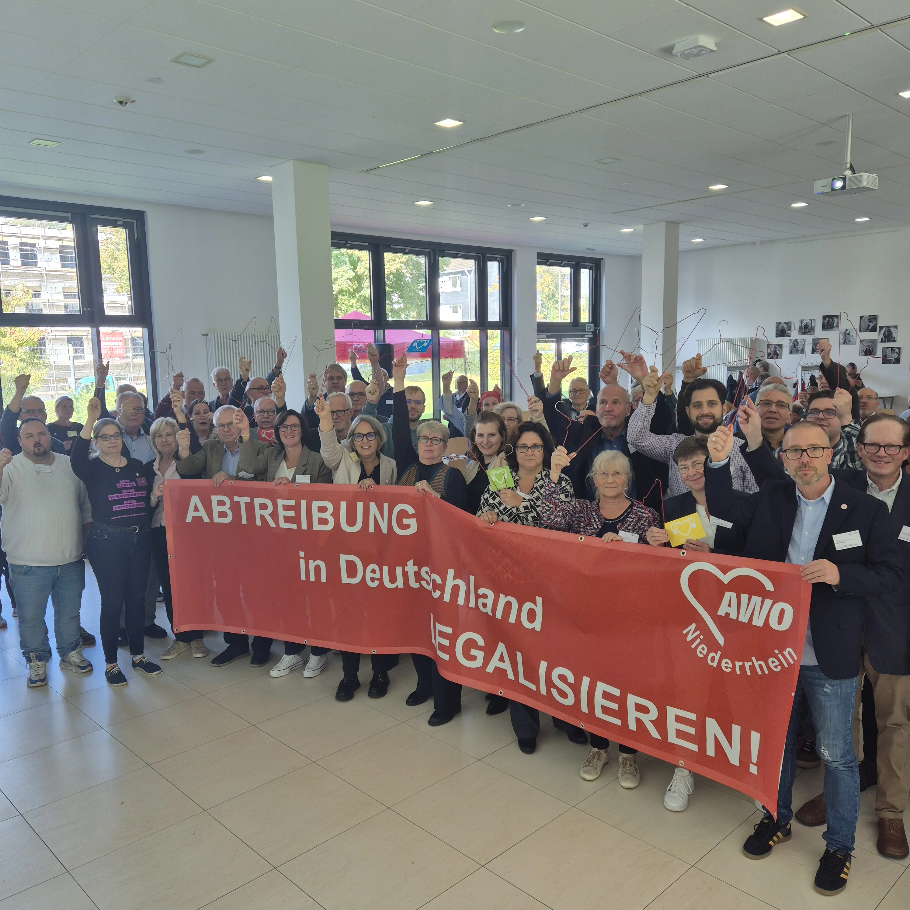 Auf dem Foto ist eine Gruppe von Menschen zu sehen, die sich in einem Innenraum versammelt haben und Banner halten. Ein großes rotes Banner trägt die Aufschrift “ABTREIBUNG in Deutschland LEGALISIEREN.” Auf dem Banner ist das Logo der AWO zu sehen.