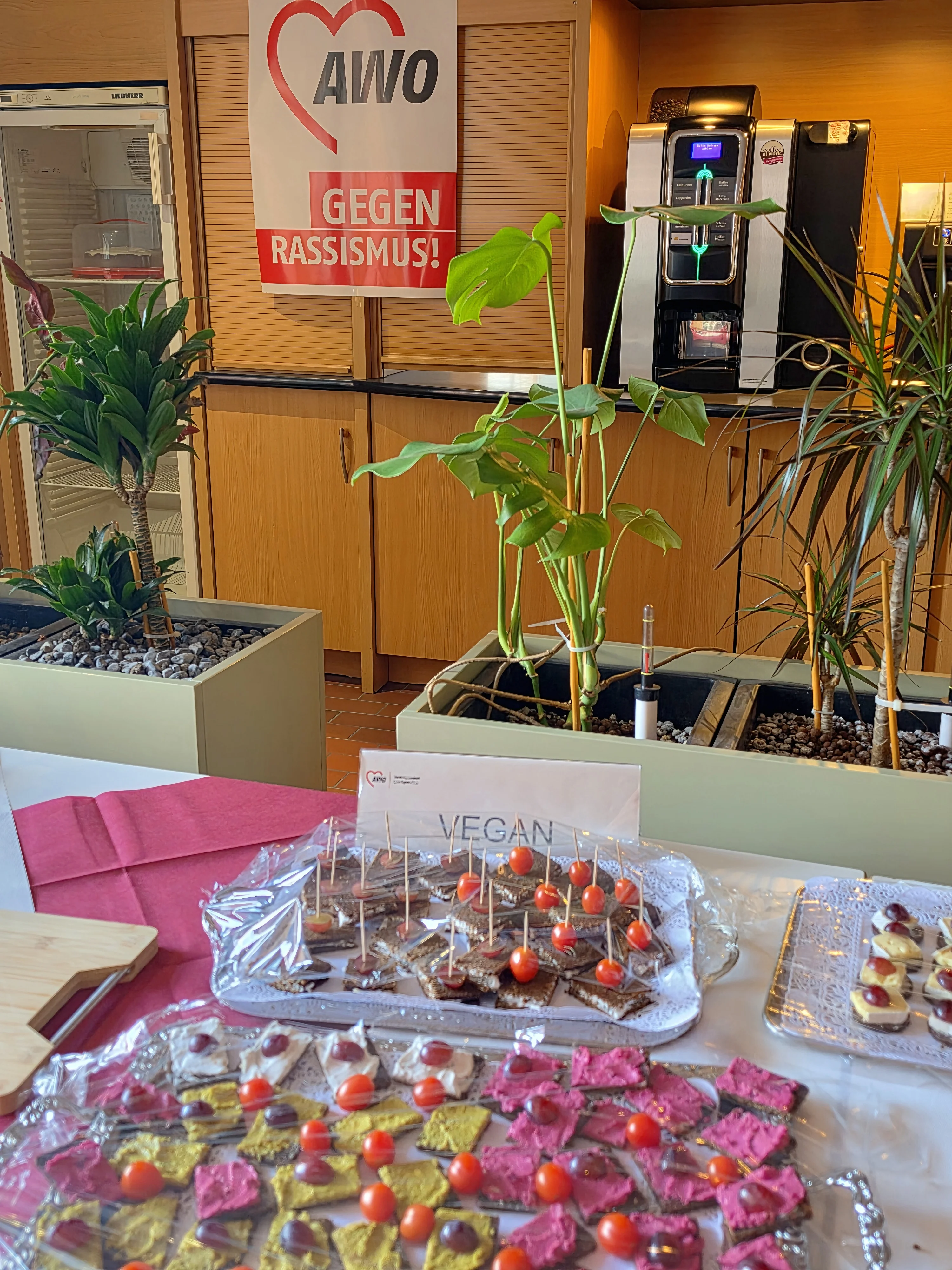 Vorne ist ein Buffet mit kleinen veganen Häppchen aufgebaut, dahinter sieht man eine Theke mit Kaffeemaschine, Pflanzen und einem AWO‑Plakat mit der Aufschrift „Gegen Rassismus!“.