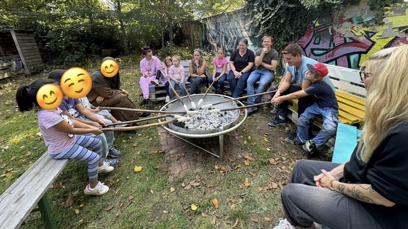 Auf dem Bild ist eine Gruppe von Erwachsenen und Kindern zu sehen, die in einem Garten um eine Feuerstelle versammelt sind. Sie sitzen auf Bänken und halten lange Stöcke, an deren Enden sie Teig über der Glut rösten. Die Stimmung wirkt entspannt und gemeinschaftlich.