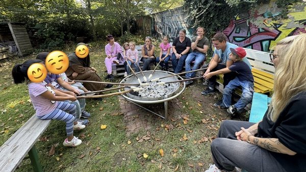 Auf dem Bild ist eine Gruppe von Erwachsenen und Kindern zu sehen, die in einem Garten um eine Feuerstelle versammelt sind. Sie sitzen auf Bänken und halten lange Stöcke, an deren Enden sie Teig über der Glut rösten. Die Stimmung wirkt entspannt und gemeinschaftlich.