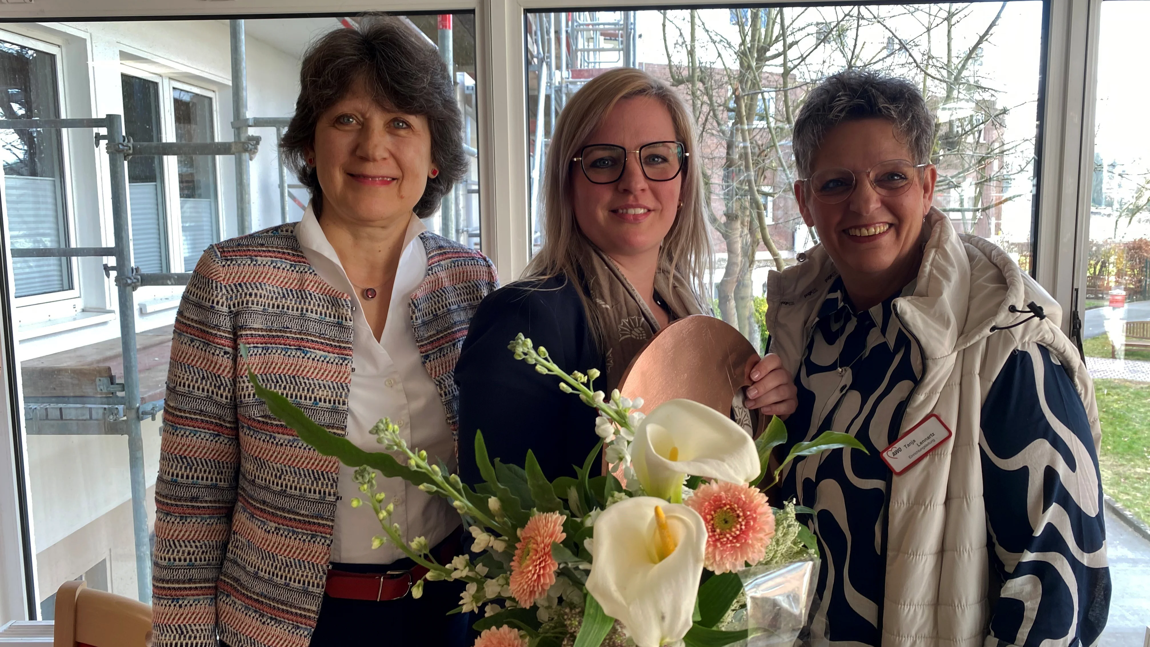 Das Foto zeigt drei Frauen, die in einem hellen Raum vor großen Fenstern stehen. Eine von ihnen hält einen Blumenstrauß mit weißen Calla-Lilien und rosa Gerbera sowie eine runde, bronzefarbene Platte. Alle drei Frauen lächeln in die Kamera. Sie tragen unterschiedliche Outfits, darunter eine gemusterte Jacke, eine dunkle Bluse mit Schal und eine Weste mit einem Namensschild. Im Hintergrund sind Fenster mit Blick auf eine Grünfläche sowie ein Gebäude mit Gerüst zu sehen.
