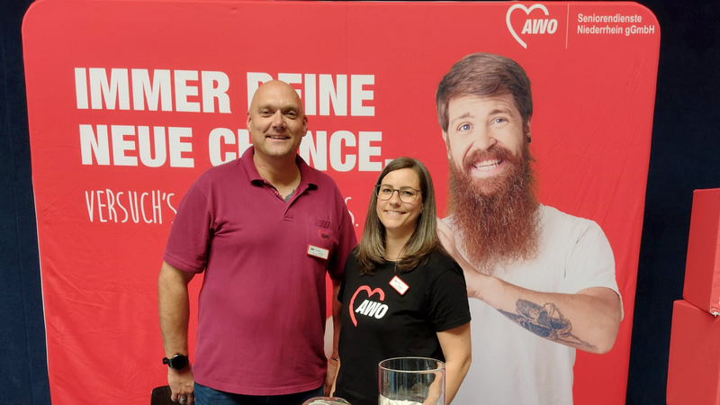 Auf dem Foto ist ein Informationsstand der AWO Seniorendienste Niederrhein gGmbH zu sehen, vermutlich im Rahmen einer Pflegemesse oder Berufsmesse (z. B. für Pflegeberufe oder soziale Berufe). Folgendes ist auf dem Bild erkennbar: Zwei Personen (eine Frau und ein Mann) stehen hinter einem roten Infotresen mit dem AWO-Logo und dem Schriftzug: „AWO Seniorendienste Niederrhein gGmbH – sozial.politisch.gestaltend.“ Im Hintergrund steht ein großer roter AWO-Werbebanner mit einem auffälligen Spruch: „IMMER DEINE NEUE CHANCE – Versuch’s mit uns.“ und einem lächelnden Mann mit Vollbart und Tattoos – ein modernes, sympathisches Image. Auf dem Tresen befinden sich: Flyer, Broschüren und Info-Materialien zur AWO Zwei Gläser mit Giveaways, vermutlich mit kleinen Gegenständen wie Buttons oder Bonbons Visitenkarten und weitere Kleinteile in kleinen Boxen Der Veranstaltungsort scheint eine Sporthalle zu sein (erkennbar an den Sportlinien auf dem Boden und der typischen Wandverkleidung).