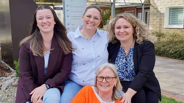  Was ist auf dem Bild zu sehen? Anhang 2026-Abschluss-der-Kita-Management-Fortbildung.jpg Auf dem Bild sitzen vier Personen draußen auf einer Bank oder einem niedrigen Sitzmöbel in einem Garten- bzw. Hofbereich. Drei Personen sitzen hinten nebeneinander, die vordere Person sitzt quer davor. Die vordere Person trägt ein auffällig orangenes Oberteil und zeigt mit beiden Händen den Daumen nach oben. Die Personen hinten tragen eher sportlich‑schicke Alltagskleidung (z.B. Blazer, Bluse, Hemd, Jeans). Im Hintergrund sind ein Weg, Grünflächen, ein Gebäude und ein großes, senkrechtes Hinweisschild mit mehreren Wegweisern zu sehen.