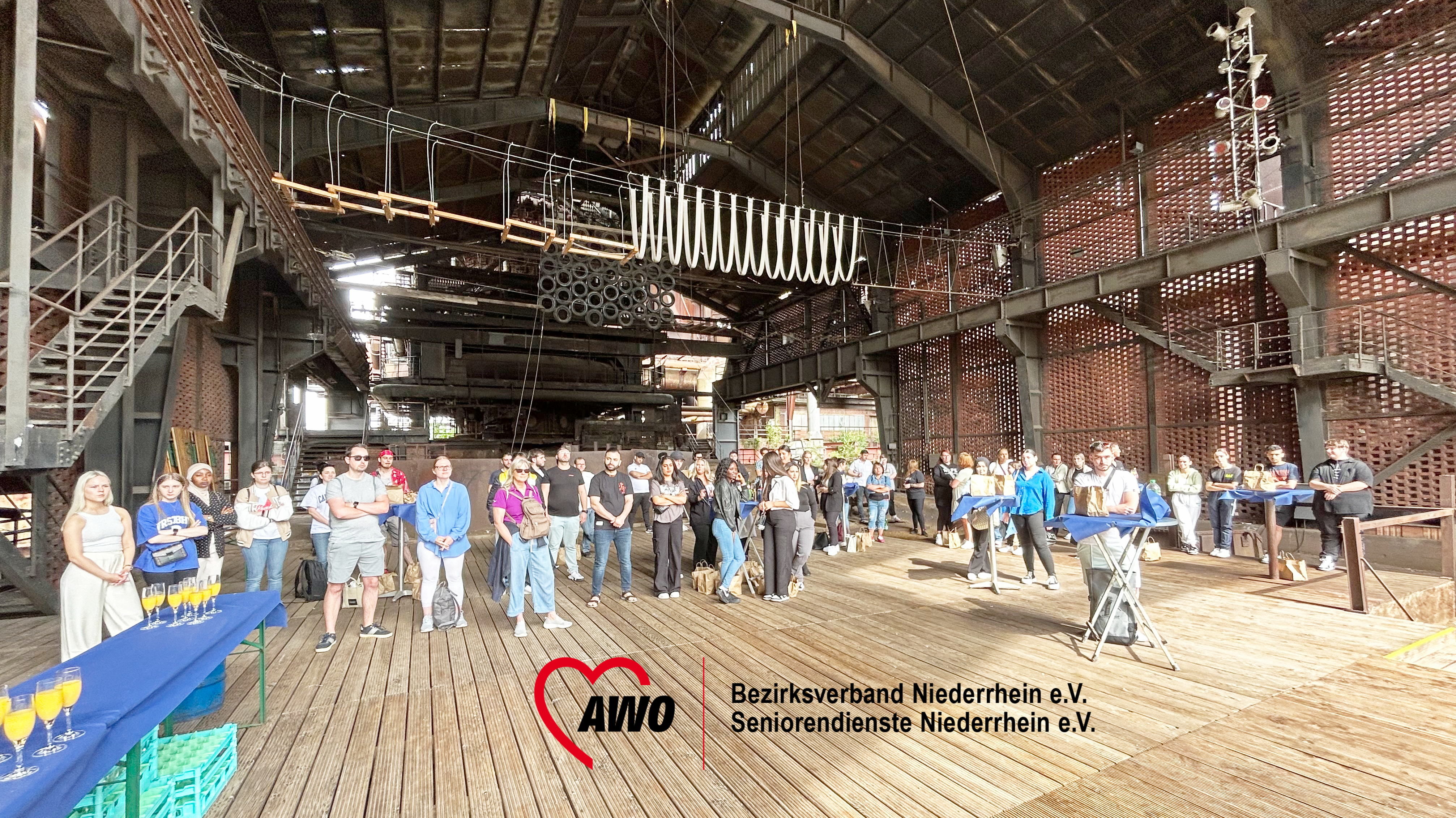 Das Foto zeigt eine Gruppe von Menschen, die sich in einer großen, industriell aussehenden Halle mit hoher Decke und Metallstrukturen versammelt haben. Die Halle hat mehrere Ebenen mit Gehwegen und wird von Tageslicht beleuchtet, das durch die Fenster fällt. An den Wänden und der Decke verlaufen Rohre, was darauf hindeutet, dass es sich um eine alte Fabrik oder Anlage handeln könnte. Im Vordergrund befindet sich ein Banner mit dem Text “AWO Bezirksverband Niederrhein e.V. Seniorendienste Niederrhein e.V.”,