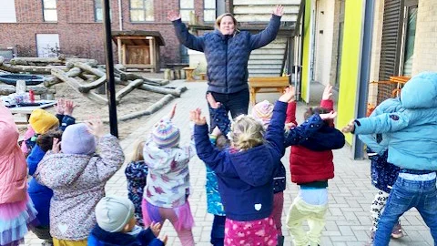 Auf dem Foto sind mehrere Kinder und eine erwachsene Person im Freien zu sehen. Die Kinder tragen warme Kleidung, darunter Mützen, Jacken und Handschuhe, was auf kaltes Wetter hindeutet. Die erwachsene Person, vermutlich eine Erzieherin oder Lehrerin, steht mit erhobenen Armen in der Mitte und scheint eine Bewegung oder Übung anzuleiten. Die Kinder stehen in einem Kreis um sie herum und machen die Bewegung nach. Der Hintergrund zeigt ein modernes Gebäude mit einer Treppe und einem Spielbereich mit Holzstrukturen.