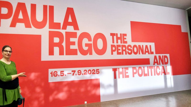 Auf dem Foto ist eine Frau zu sehen, die vor einer großen roten und weißen Wandinstallation steht. Auf der Wand ist in großen roten Buchstaben "PAULA REGO" zu lesen, gefolgt von kleinerem Text, der die Ausstellung als "THE PERSONAL AND THE POLITICAL" beschreibt. Auch das Datum der Ausstellung, "16.5.–7.9.2025", ist angegeben.