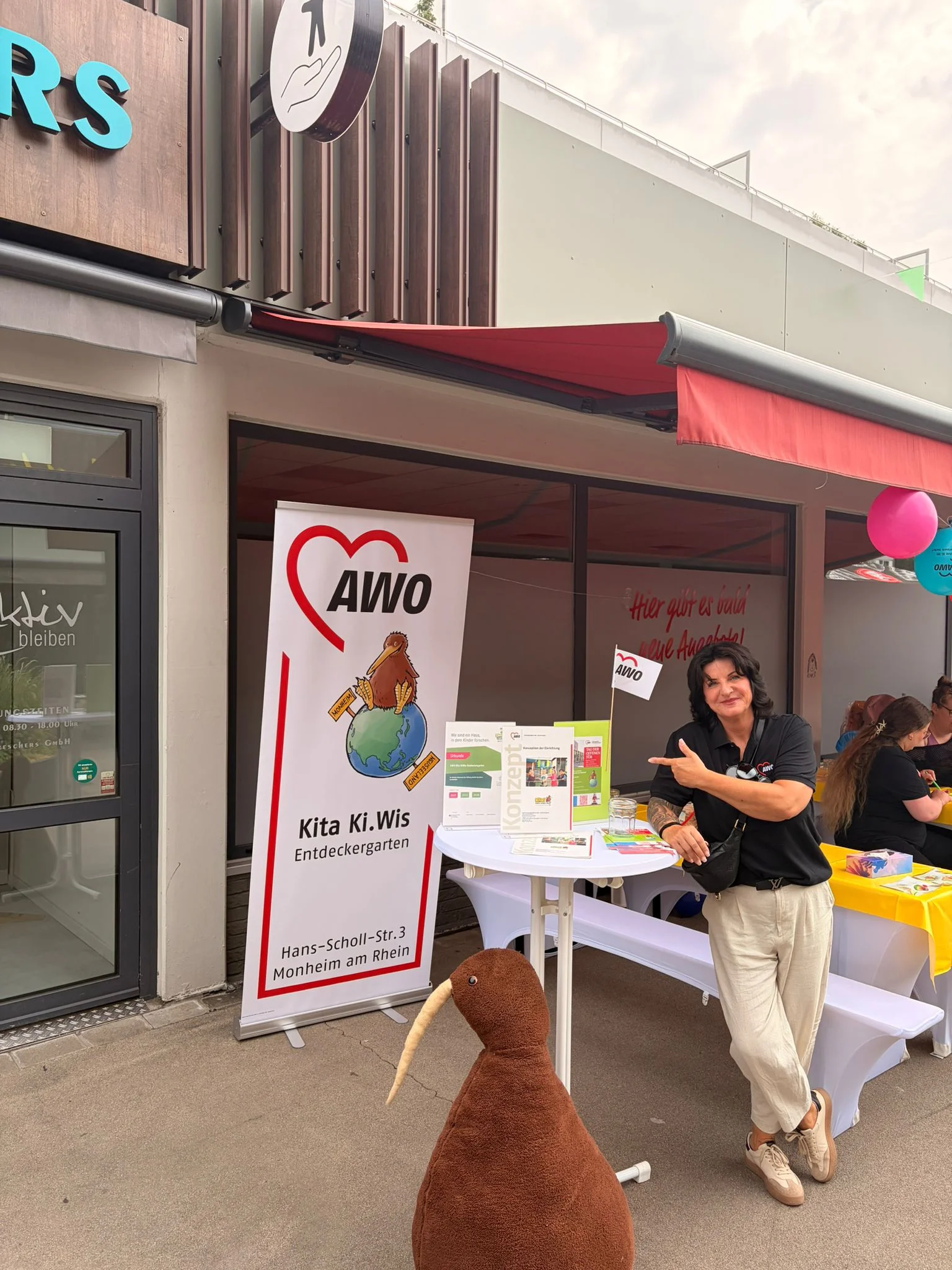 Auf dem Foto ist ein Infostand der AWO vor einem Gebäude zu sehen. Links steht ein roll‑up mit AWO‑Logo und der Aufschrift „Kita Ki.Wis Entdeckergarten – Hans‑Scholl‑Str. 3, Monheim am Rhein“, daneben ein Stehtisch mit Infomaterial und Flyern, an dem eine Person steht und auf die Unterlagen zeigt. Im Vordergrund steht ein großes braunes Stofftier in Kiwi‑Vogel‑Form, im Hintergrund sitzen weitere Personen an einem gelb gedeckten Tisch, über dem bunte Luftballons hängen.