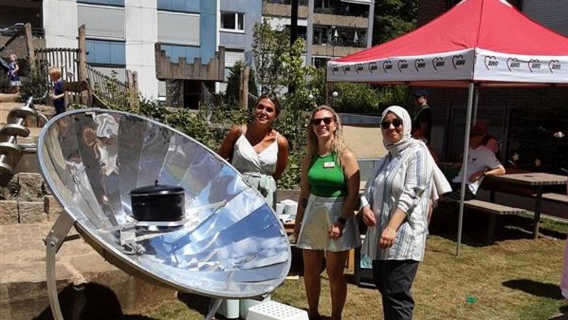 Auf dem Foto sind drei Frauen im Freien zu sehen, die vor einer großen Solarkochplatte mit einem Topf darauf stehen. Die Frau in der Mitte trägt ein grünes Oberteil und eine helle Hose, die Frau links ein helles Oberteil und eine dunkle Hose, und die Frau rechts eine gestreifte Bluse und eine dunkle Hose mit einem Kopftuch. Im Hintergrund sind Wohngebäude, Bäume und ein rotes Zelt zu erkennen. Der Boden ist größtenteils mit Gras bedeckt. Es scheint ein sonniger Tag zu sein.