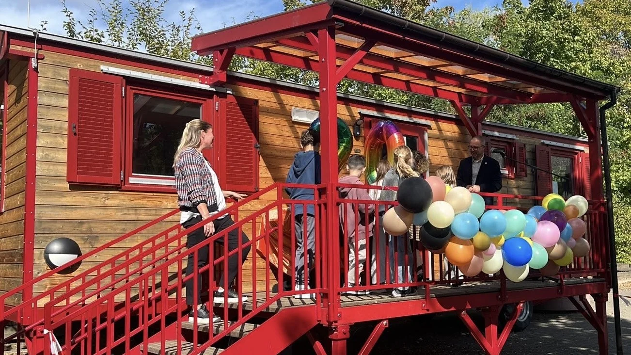 Auf dem Foto ist eine Szene im Freien vor einem Wagen oder einem kleinen, holzverkleideten Gebäude zu sehen. Es scheint sich um eine Art Veranstaltung oder kleine Feier zu handeln, denn: Das Gebäude hat eine auffällige, rot gestrichene Treppe und eine überdachte Veranda. Die Veranda ist mit einem großen Bündel von bunten Ballons (in verschiedenen Pastell- und dunkleren Tönen) geschmückt. Mehrere Personen sind auf der Treppe und der Veranda versammelt, darunter eine erwachsene Frau auf der Treppe, ein erwachsener Mann auf der Veranda und mehrere Kinder. Im Fenster über der Veranda sind auch große, folienartige Zahlen-Ballons zu erkennen, was auf einen Geburtstag oder ein Jubiläum hindeutet. Die Umgebung besteht aus Bäumen und Grünzeug im Hintergrund, und der Himmel ist blau mit Sonnenschein.