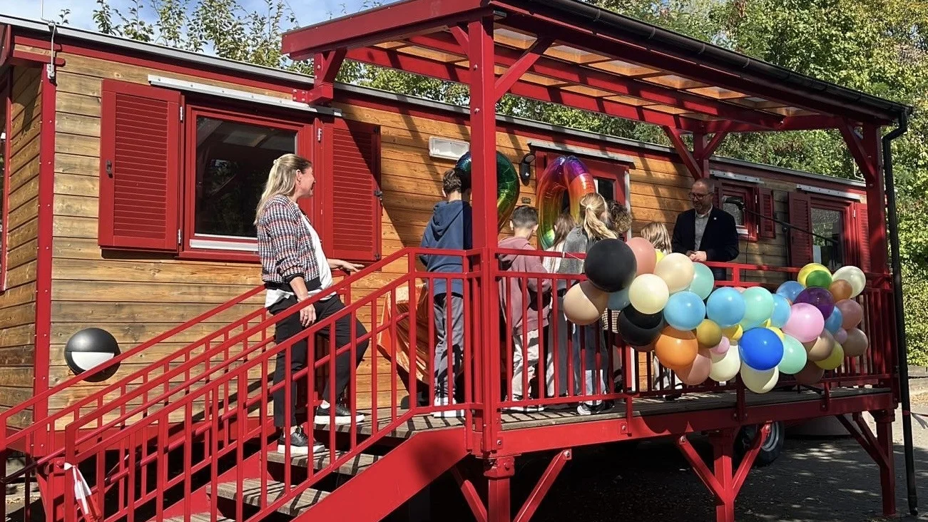 Auf dem Foto ist eine Szene im Freien vor einem Wagen oder einem kleinen, holzverkleideten Gebäude zu sehen. Es scheint sich um eine Art Veranstaltung oder kleine Feier zu handeln, denn: Das Gebäude hat eine auffällige, rot gestrichene Treppe und eine überdachte Veranda. Die Veranda ist mit einem großen Bündel von bunten Ballons (in verschiedenen Pastell- und dunkleren Tönen) geschmückt. Mehrere Personen sind auf der Treppe und der Veranda versammelt, darunter eine erwachsene Frau auf der Treppe, ein erwachsener Mann auf der Veranda und mehrere Kinder. Im Fenster über der Veranda sind auch große, folienartige Zahlen-Ballons zu erkennen, was auf einen Geburtstag oder ein Jubiläum hindeutet. Die Umgebung besteht aus Bäumen und Grünzeug im Hintergrund, und der Himmel ist blau mit Sonnenschein.