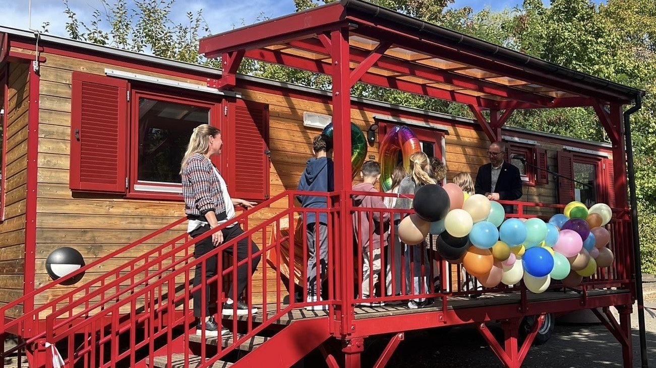  Auf dem Foto ist eine Szene im Freien vor einem Wagen oder einem kleinen, holzverkleideten Gebäude zu sehen. Es scheint sich um eine Art Veranstaltung oder kleine Feier zu handeln, denn: Das Gebäude hat eine auffällige, rot gestrichene Treppe und eine überdachte Veranda. Die Veranda ist mit einem großen Bündel von bunten Ballons (in verschiedenen Pastell- und dunkleren Tönen) geschmückt. Mehrere Personen sind auf der Treppe und der Veranda versammelt, darunter eine erwachsene Frau auf der Treppe, ein erwachsener Mann auf der Veranda und mehrere Kinder. Im Fenster über der Veranda sind auch große, folienartige Zahlen-Ballons zu erkennen, was auf einen Geburtstag oder ein Jubiläum hindeutet. Die Umgebung besteht aus Bäumen und Grünzeug im Hintergrund, und der Himmel ist blau mit Sonnenschein.