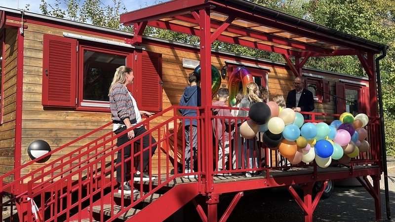  Auf dem Foto ist eine Szene im Freien vor einem Wagen oder einem kleinen, holzverkleideten Gebäude zu sehen. Es scheint sich um eine Art Veranstaltung oder kleine Feier zu handeln, denn: Das Gebäude hat eine auffällige, rot gestrichene Treppe und eine überdachte Veranda. Die Veranda ist mit einem großen Bündel von bunten Ballons (in verschiedenen Pastell- und dunkleren Tönen) geschmückt. Mehrere Personen sind auf der Treppe und der Veranda versammelt, darunter eine erwachsene Frau auf der Treppe, ein erwachsener Mann auf der Veranda und mehrere Kinder. Im Fenster über der Veranda sind auch große, folienartige Zahlen-Ballons zu erkennen, was auf einen Geburtstag oder ein Jubiläum hindeutet. Die Umgebung besteht aus Bäumen und Grünzeug im Hintergrund, und der Himmel ist blau mit Sonnenschein.