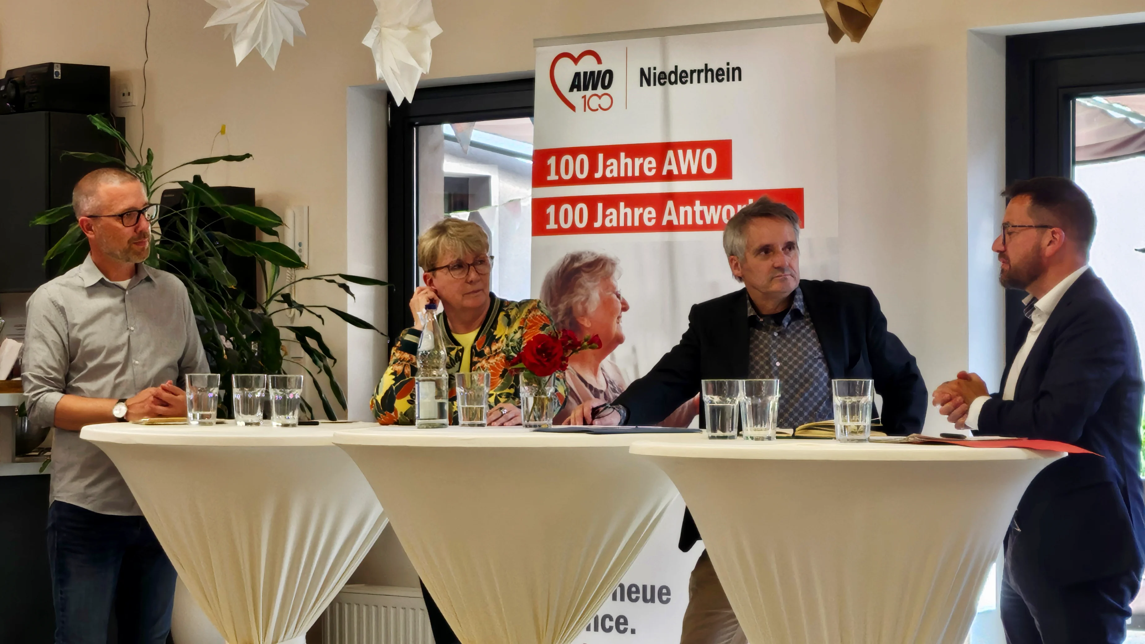 Das Foto zeigt eine Gruppe von Personen, die während einer Veranstaltung an einem Tisch stehen. Es sind vier Menschen zu sehen, die als Teilnehmende einer Podiumsdiskussion hinter einem mit einer weißen Tischdecke bedeckten Tisch stehen. Im Hintergrund befindet sich ein Banner mit dem Text “A 100 Jahre AWO. 100 Jahre Antworten” und es gibt weitere Dekorationen wie weiße Vorhänge und Blumen, die zu einer offiziellen und festlichen Atmosphäre beitragen.