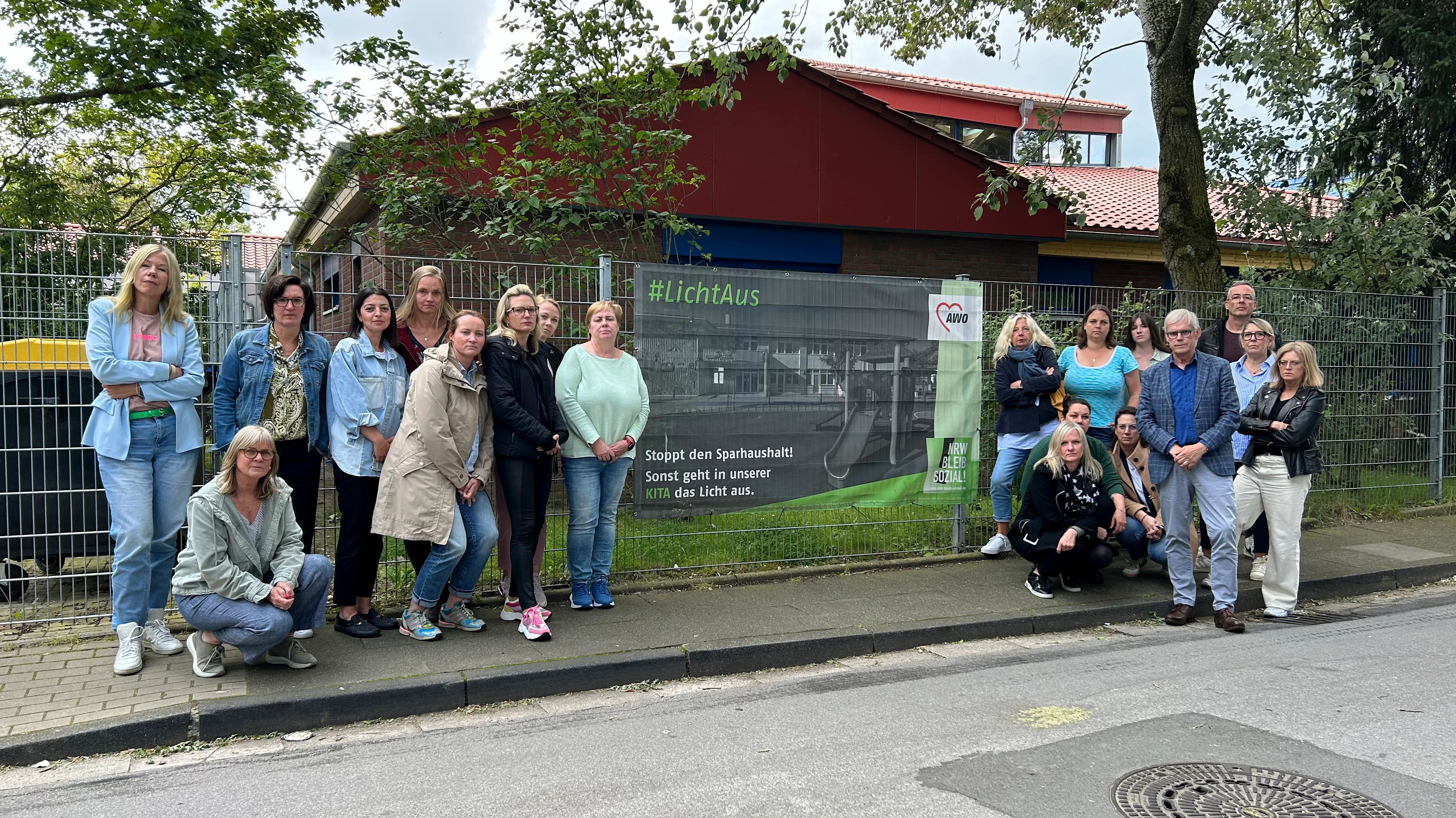 Das Foto zeigt eine Gruppe von Personen, die vor einem Zaun stehen. Hinter dem Zaun befindet sich grüne Vegetation. Im Hintergrund ist ein Gebäude mit einem roten Dach zu sehen. Ein Banner am Zaun enthält Text den: „#lichtaus. Stoppt den Sparhaushalt. Sonst geht in unserer Kita das Licht aus.“ Zudem ist auf dem schwarz-grünen Banner das Logo der AWO zu sehen.
